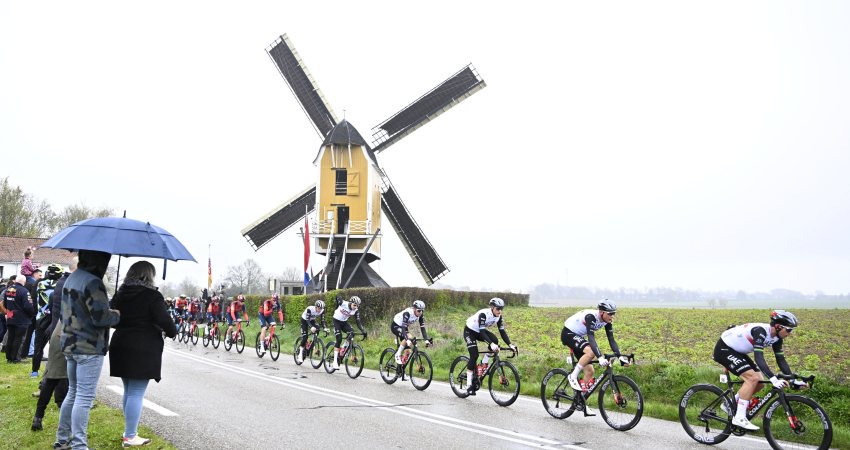 Mensen onder een paraplu kijken hoe wielrenners tijdens de Amstel Gold Race van 2023 door een regenachtig Limburgs landschap koersen. 